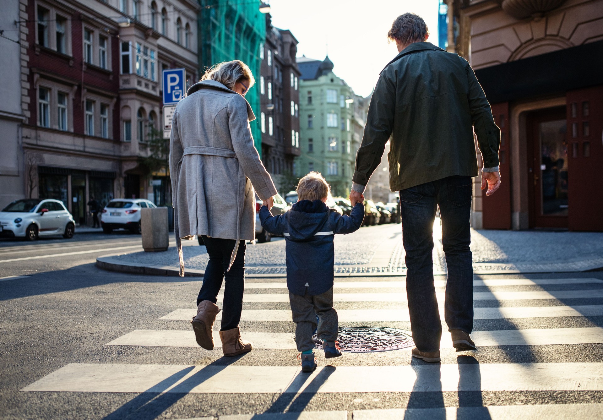 A rear view of small toddler boy with parents crossing a road outdoors in city.
